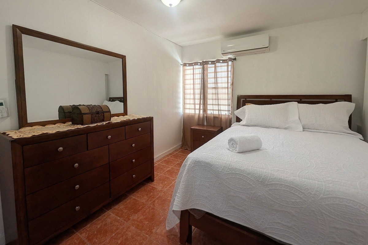 A bedroom with a wooden dresser, a bed with white bedding, a large mirror, an air conditioning unit, and a window with curtains.