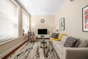 Ein Wohnzimmer mit einer Couch, einem Sessel, einem Fernseher, einer Uhr und einem gerahmten Bild an der Wand.