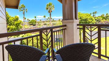 A balcony with two wicker chairs, a view of a landscaped area with palm trees, and a clear sky.