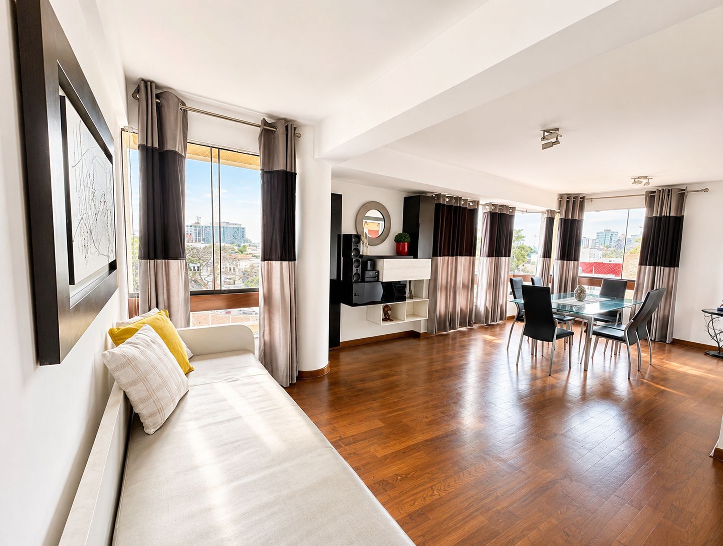 Una sala moderna con sofá, mesa de comedor y sillas, un área de cocina con refrigerador y armarios, y ventanas grandes con cortinas.