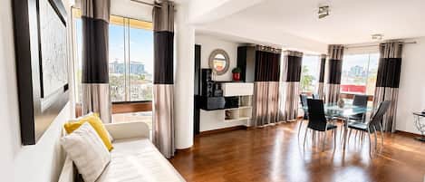 A modern living room with a sofa, dining table, and chairs, a kitchen area with a fridge and cabinets, and large windows with curtains.