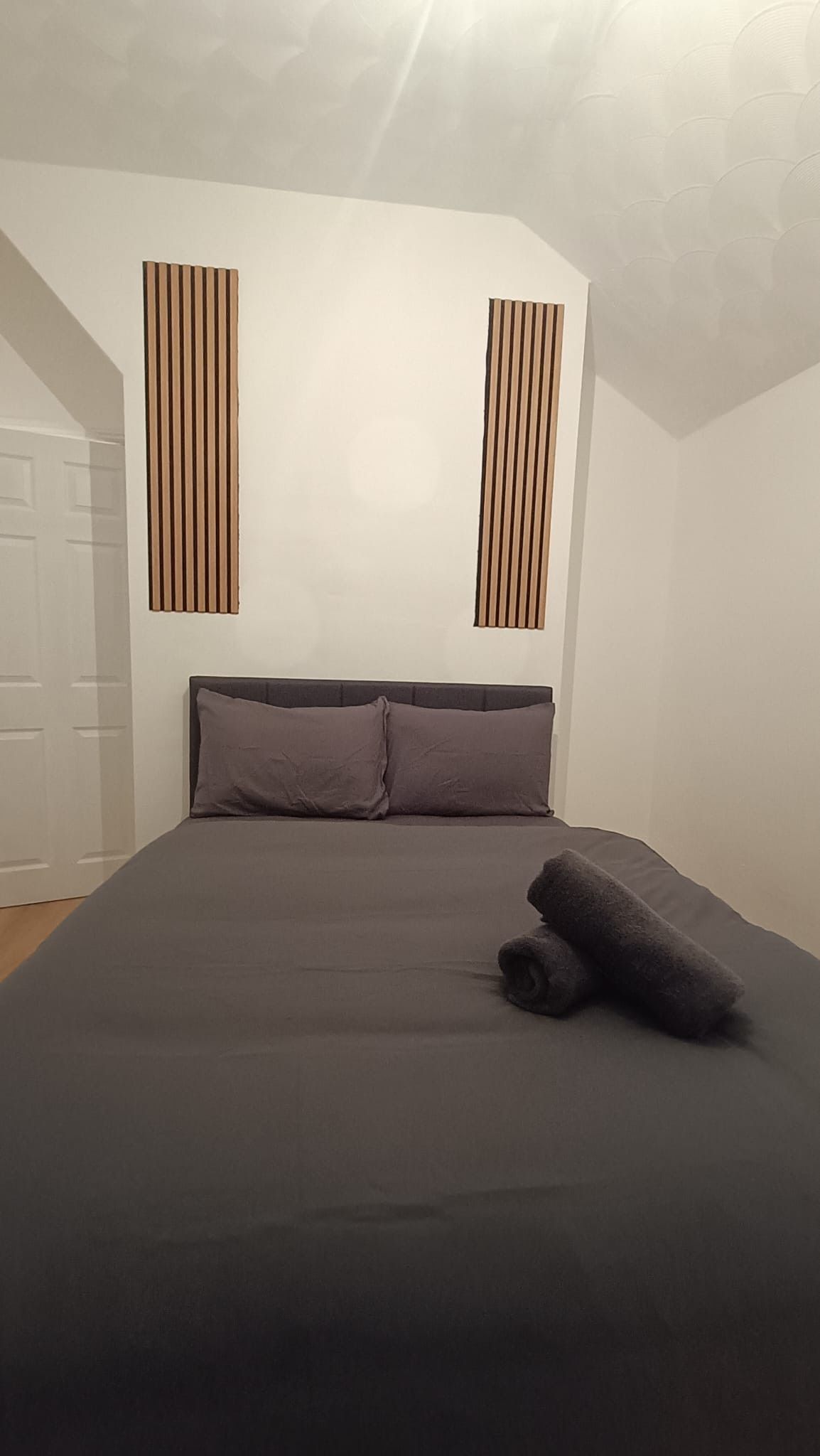 A bedroom with a bed, pillows, and a rolled-up blanket. There are two vertical wooden slats on the wall.