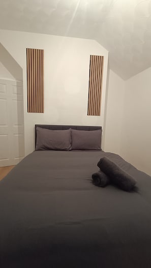 Une chambre à coucher avec un lit, des oreillers et une couverture enroulée. Il y a deux lambris de bois verticaux sur le mur.