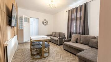 Un salon avec un canapé, une petite table, une horloge et un téléviseur fixé au mur.