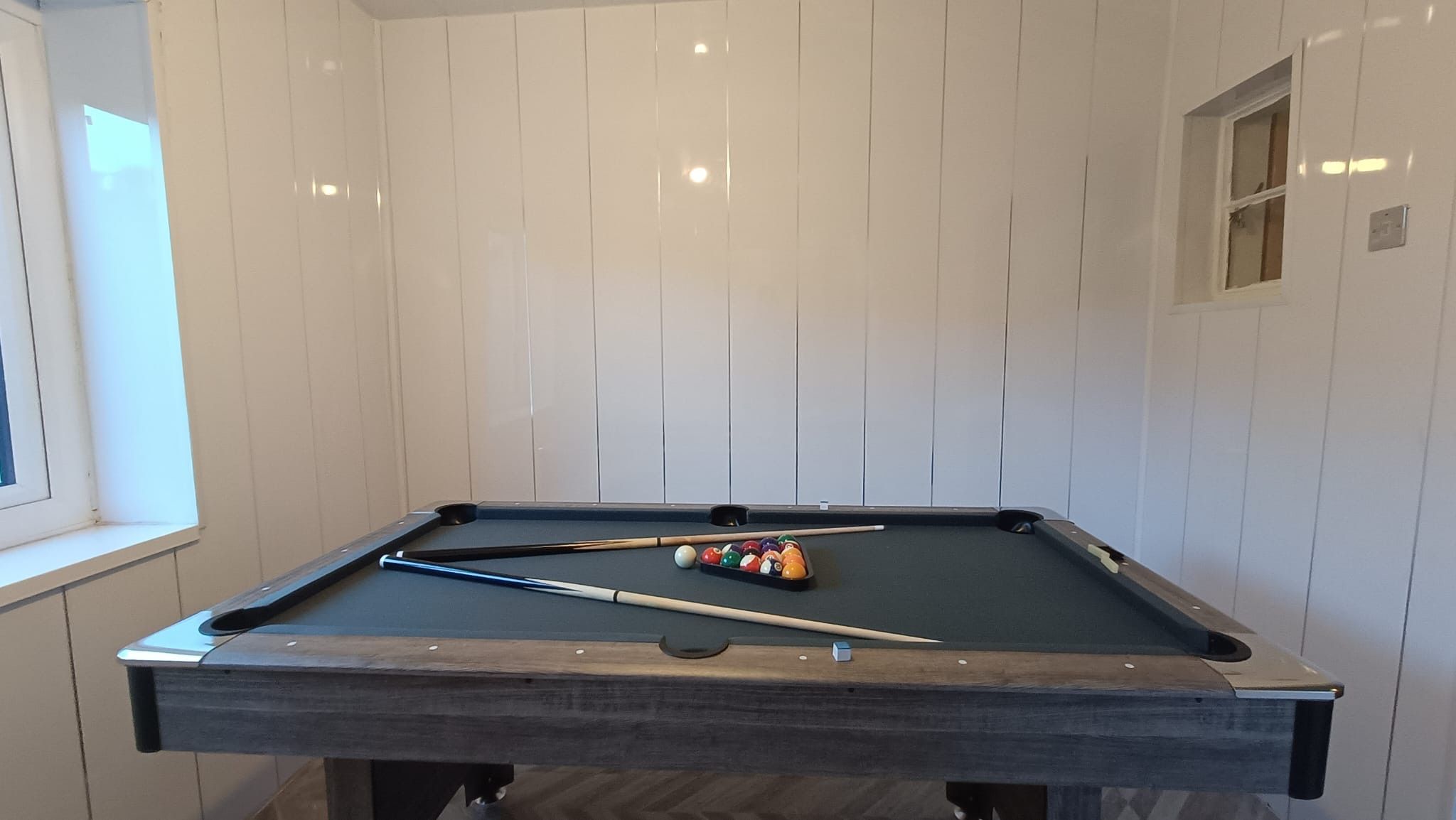 Ein Billardtisch mit Kugeln und Spielstöcken vor einer weißen Wand mit Paneelen.