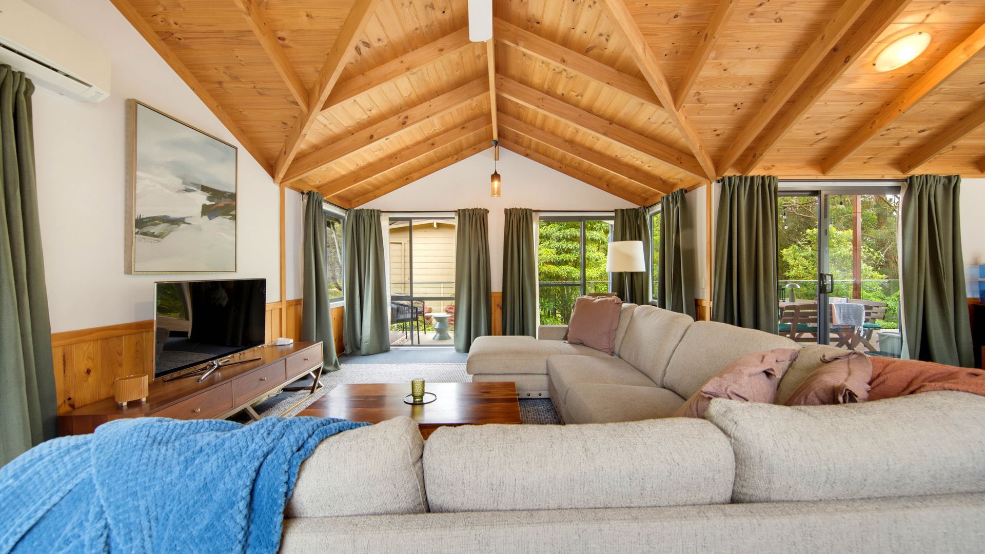 A spacious living room with a high wooden ceiling, a large sofa, a television, and a view of the outdoors through large windows.