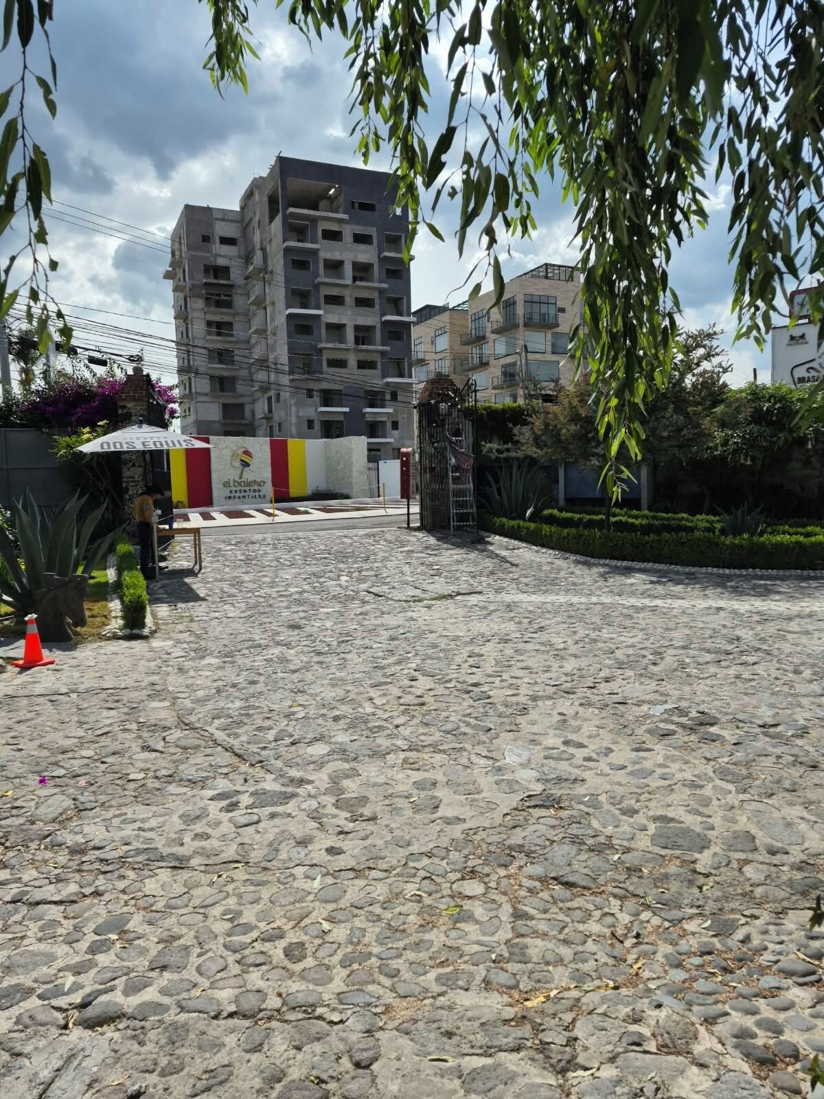 A paved courtyard with a building in the background, a sign with 'el balen' and 'BRASIL' visible, and a tree with green leaves.