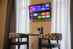 A television mounted on a wall displaying various apps, a table with a kettle, and two chairs with patterned cushions. - LUNA PLAZA HOTEL TASHKENT (Tashkent)