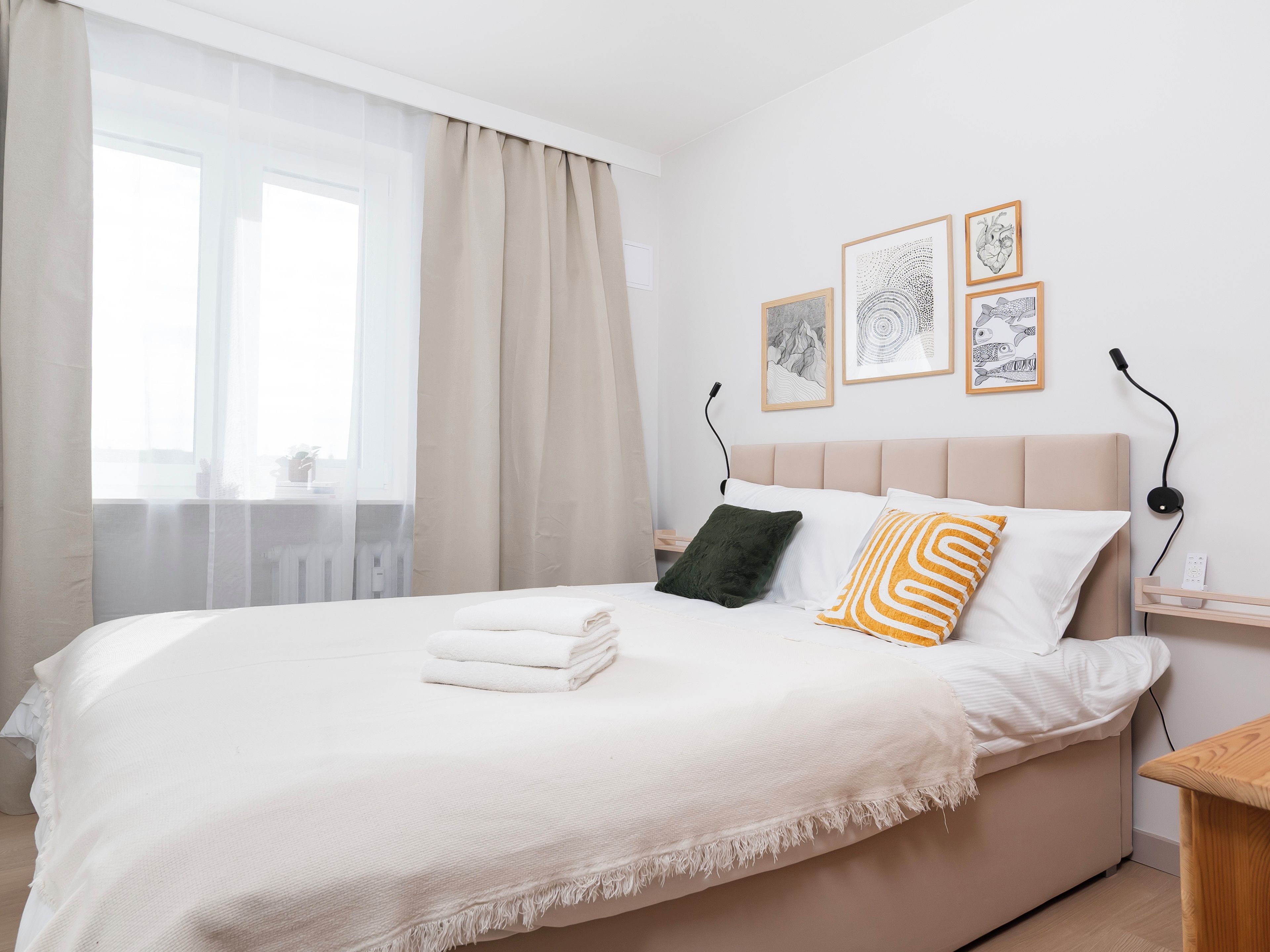 Un lit bien fait, avec une couette blanche et une serviette pliée, une table de chevet avec une lampe, et un mur orné de tableaux encadrés.