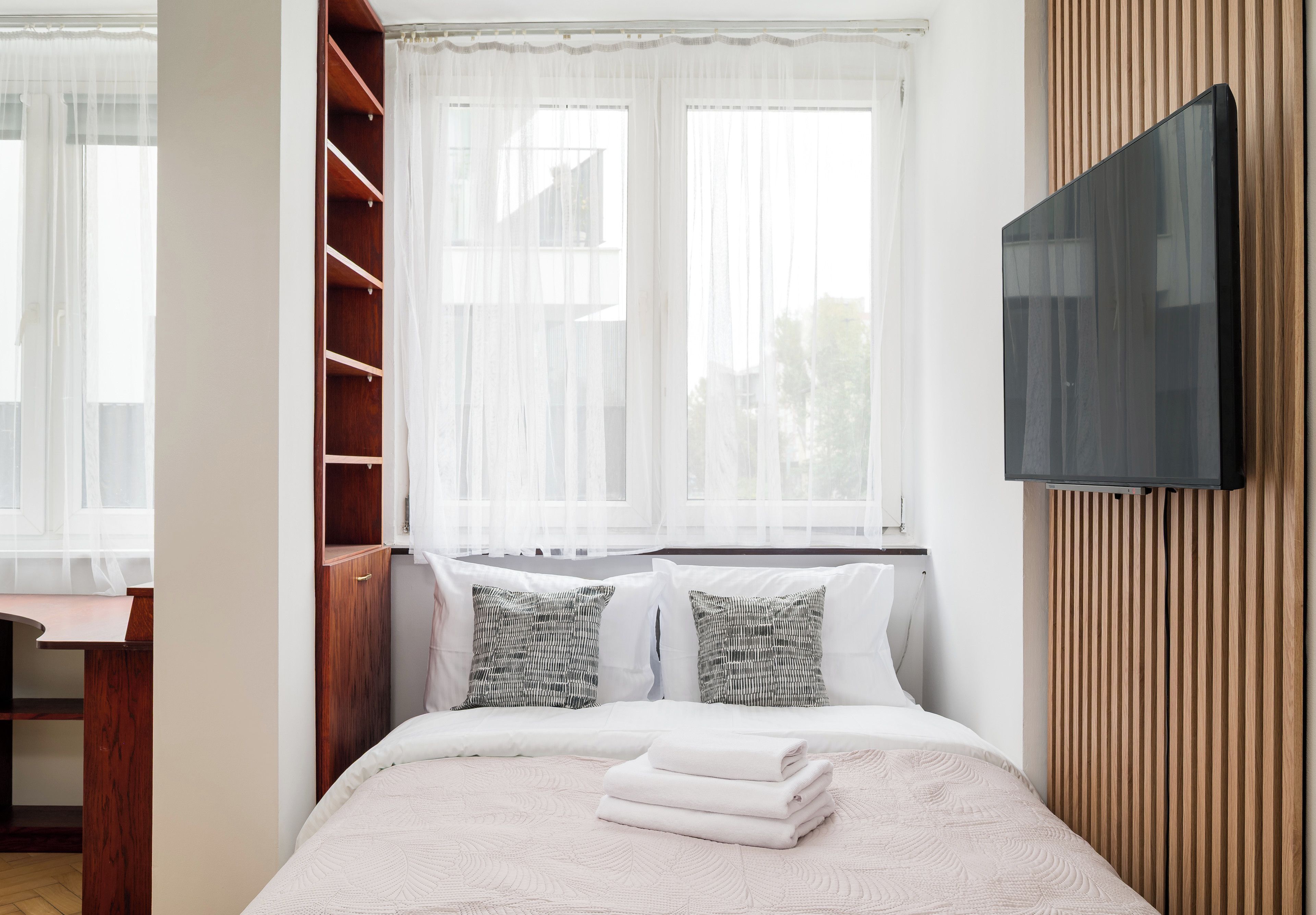 Un dormitorio con una cama, un televisor montado en la pared, un armario de madera y un escritorio.