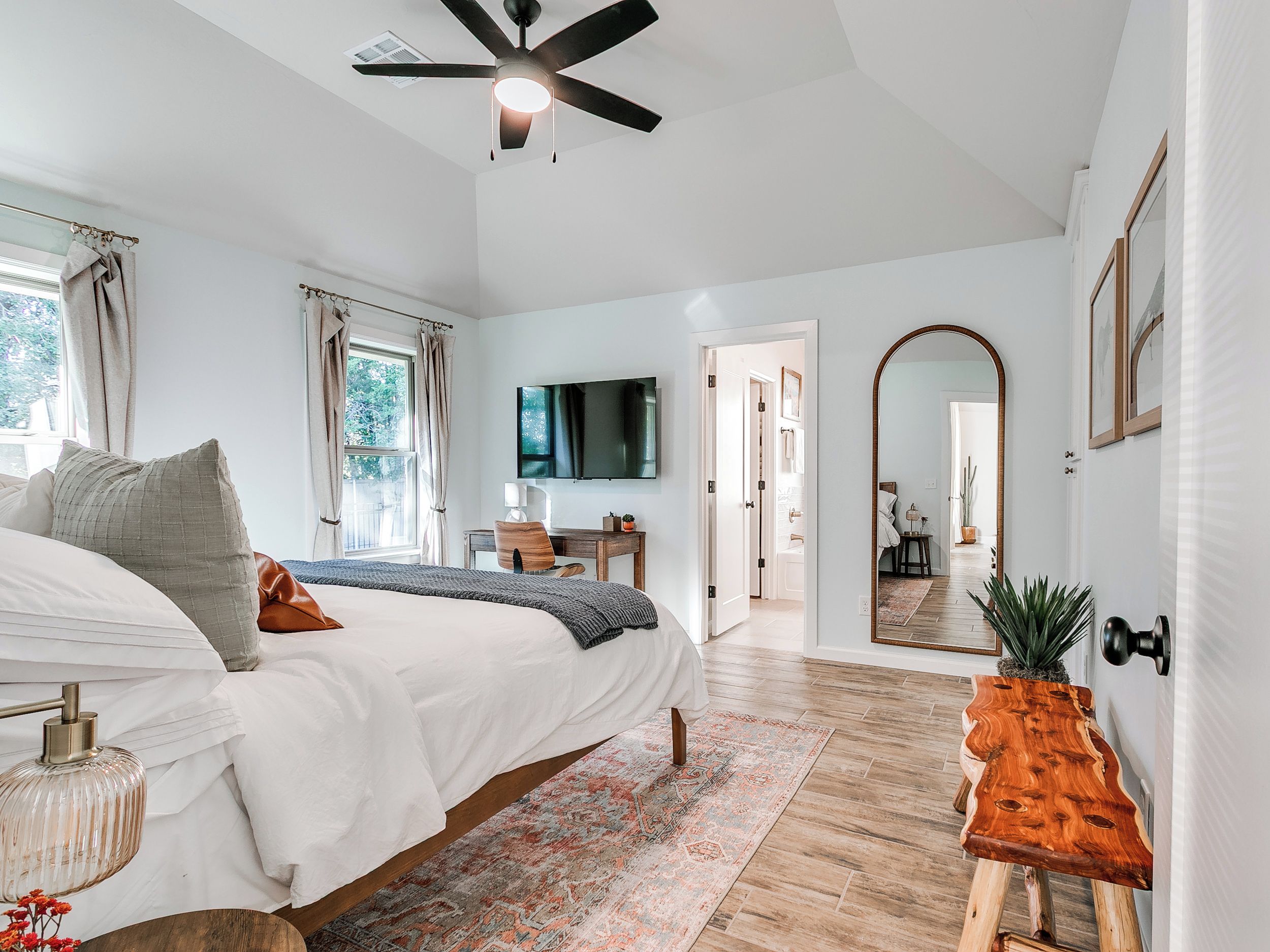 A bedroom with a bed, bedside table, and a desk with a chair. There is a ceiling fan, a TV mounted on the wall, and a large mirror.