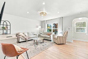 Un salon moderne avec un canapé d’angle, une table basse ronde, un fauteuil à rayures et une grande fenêtre avec des rideaux.