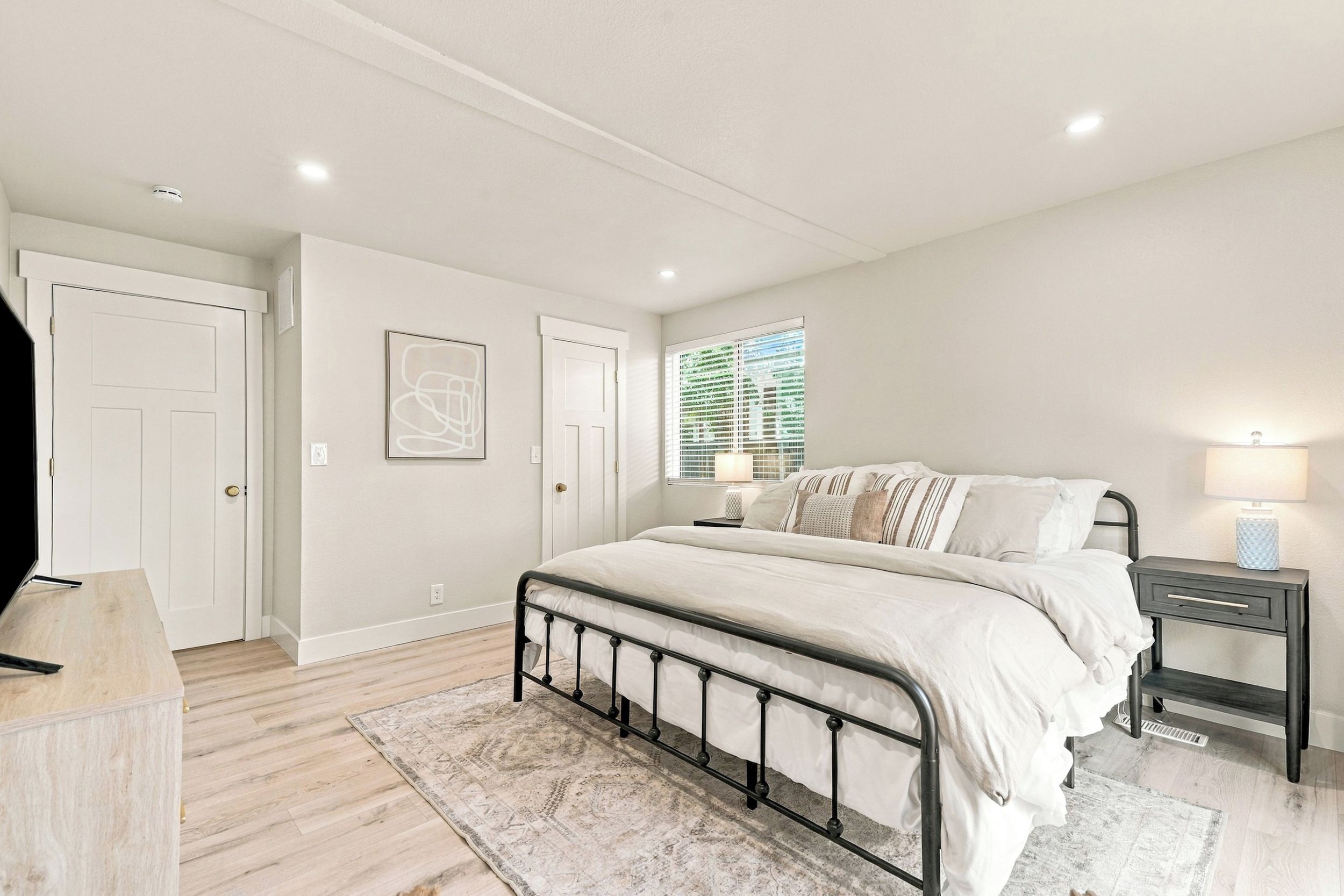 Un dormitorio con cama, mesitas de noche, una ventana con persianas y una puerta.