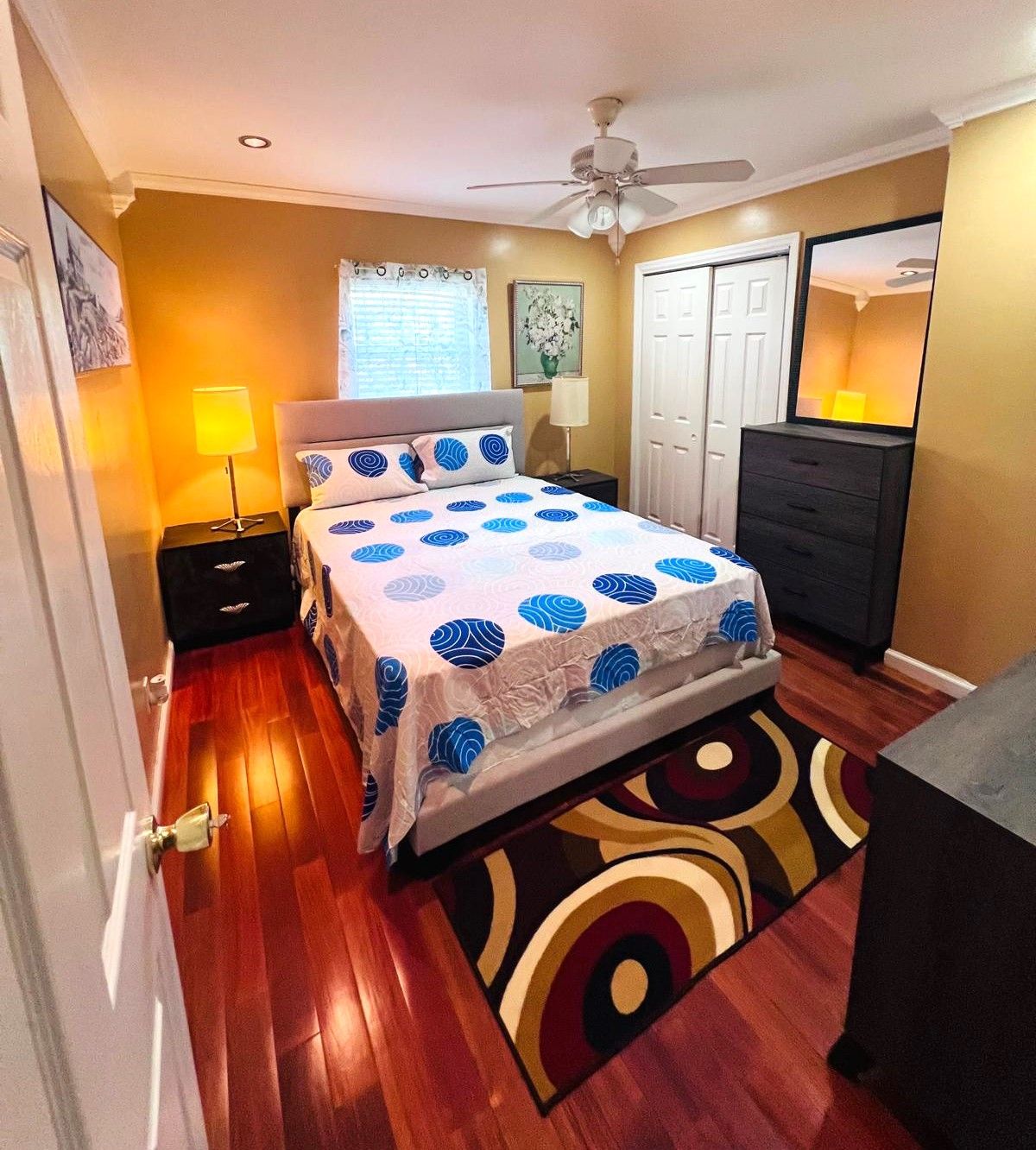 A bedroom with a bed, bedside tables, a dresser, a ceiling fan, and a window with curtains.
