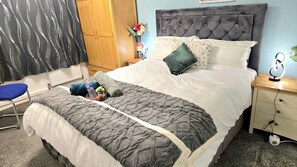 Una cama bien tendida con una manta gris texturizada, una silla azul, una mesita de noche con una lámpara y un armario de madera.