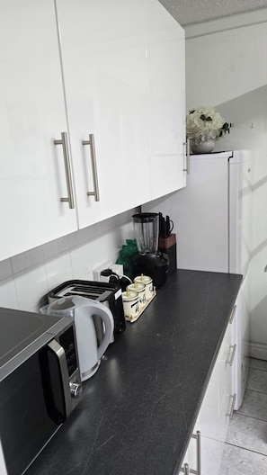 Una cocina con armarios blancos, encimera negra, tostadora, hervidor, licuadora y refrigerador.