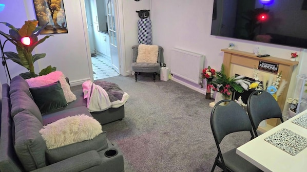 Una sala de estar con un sofá, sillones, una mesa de centro y un televisor. Hay un radiador, una chimenea y una mesa de comedor con sillas.