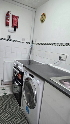 Een kleine keuken met een wasmachine, een brandblusser en een branddeken aan de muur.