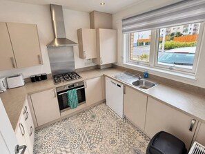 A compact kitchen with a gas stove, oven, and dishwasher, a window with a view of the street, and patterned floor tiles. - Bright,modern 4 bedroom house with free Parking (Edinburgh)