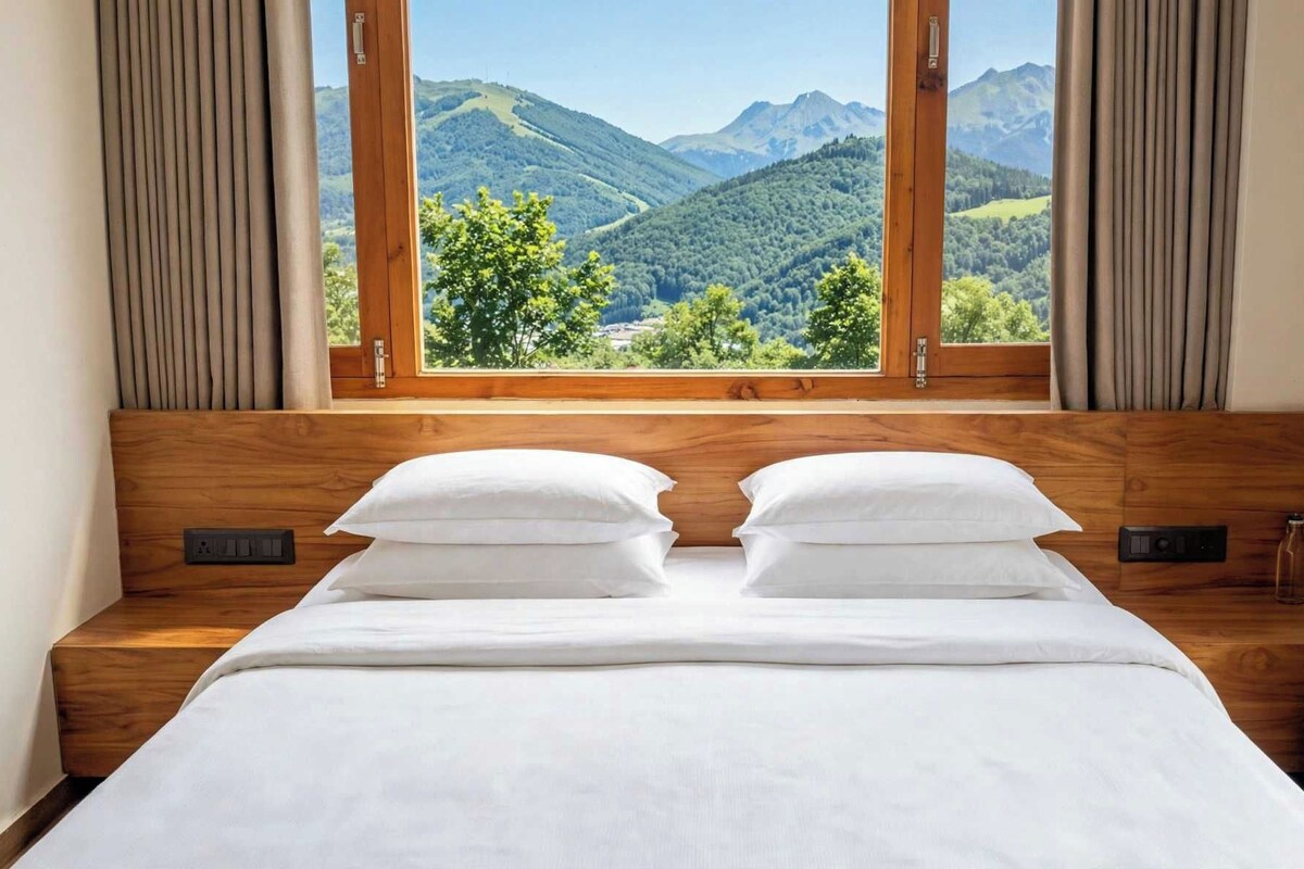 Ein Schlafzimmer mit hölzernem Kopfteil, zwei weißen Kissen, einer weißen Tagesdecke und einem Fenster mit Blick auf grüne Hügel und Berge.