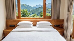 Ein Schlafzimmer mit hölzernem Kopfteil, zwei weißen Kissen, einer weißen Tagesdecke und einem Fenster mit Blick auf grüne Hügel und Berge.