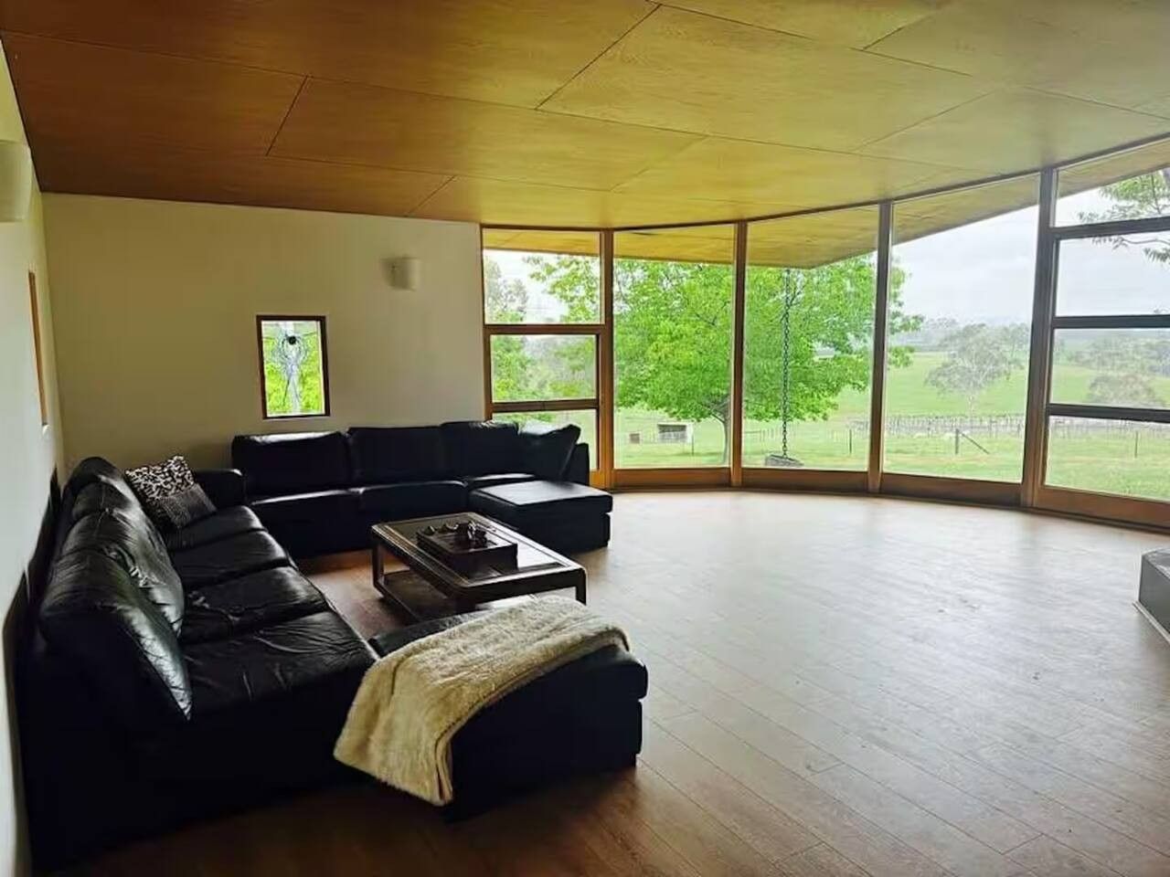 A spacious living room with a black leather sofa, a coffee table, and large windows offering a view of greenery outside.