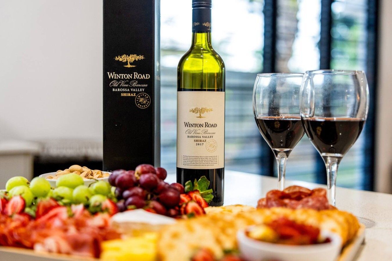 A bottle of Winton Road wine, two glasses of red wine, and a selection of cheeses and fruits on a table.