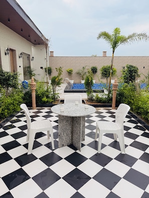 Une terrasse extérieure au sol carrelé, avec une table ronde et des chaises blanches, entourée de plantes en pot et d’une piscine.