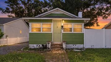 Une maison de plain-pied à l’extérieur vert, avec une clôture blanche en bois et un chemin de briques menant à la porte d’entrée.