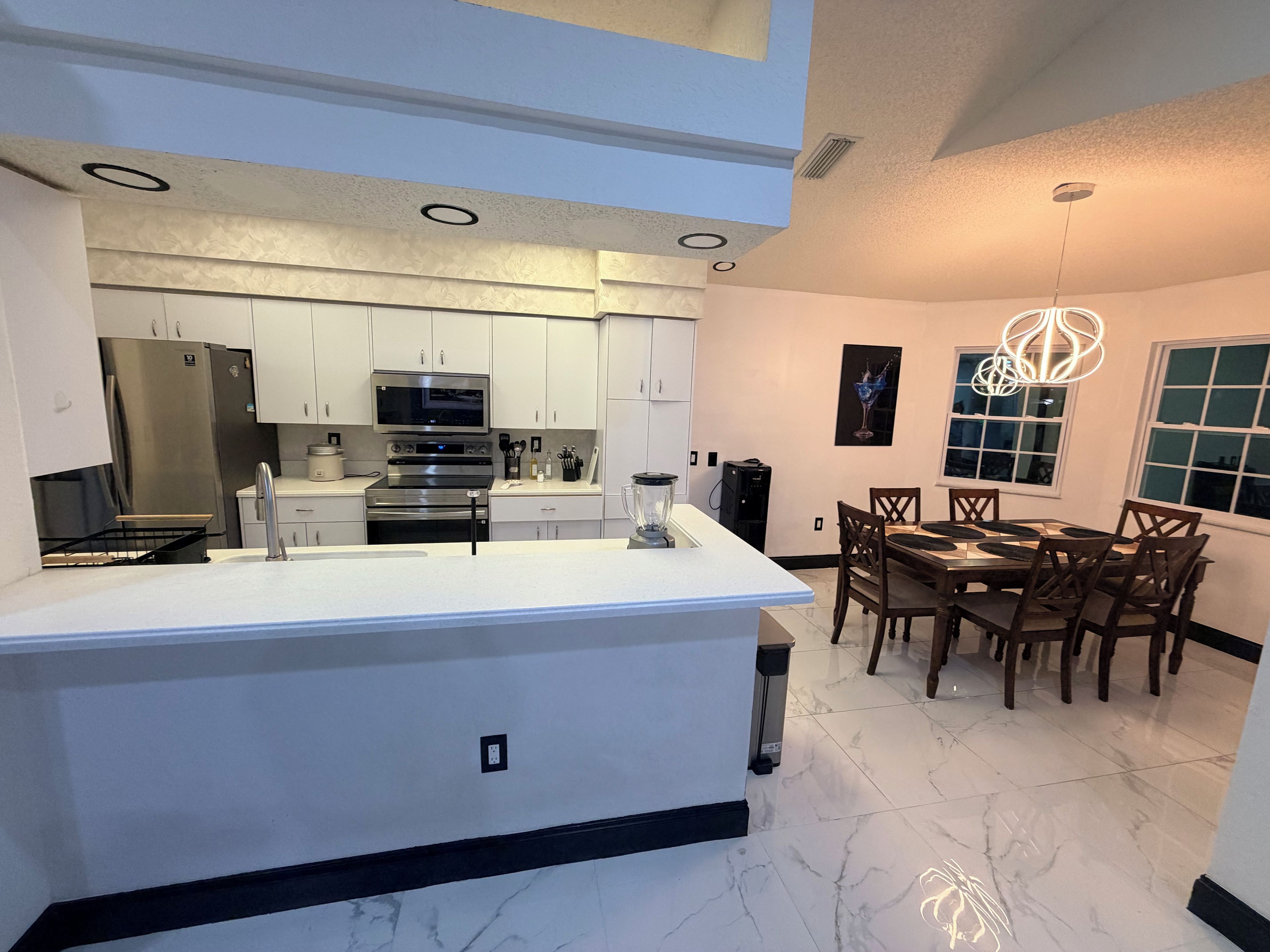 A modern kitchen with a central island, white cabinetry, stainless steel appliances, and a dining area with wooden chairs and a table.