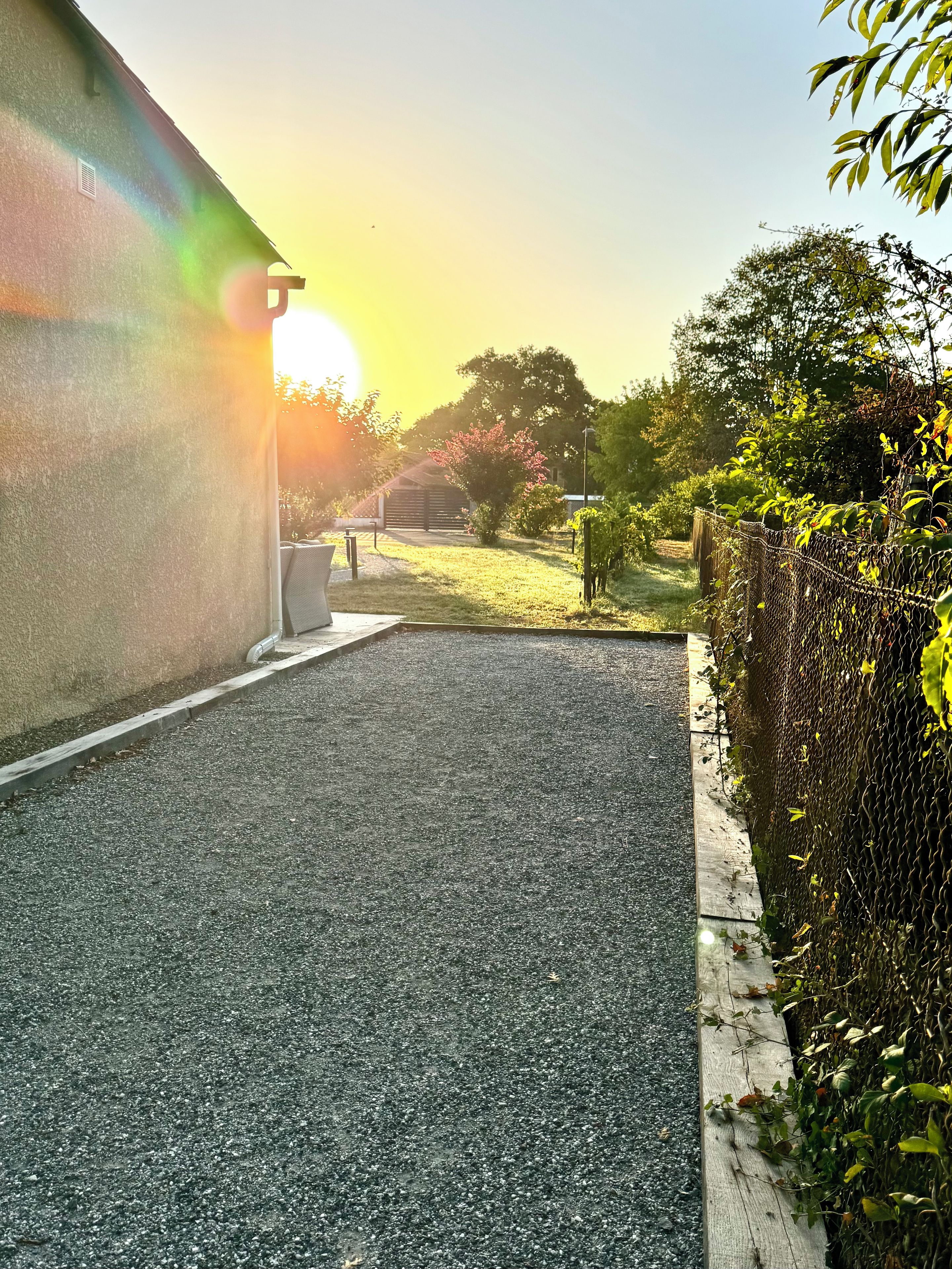 A paved driveway leading to a building with a sloped roof, surrounded by greenery and a chain-link fence.