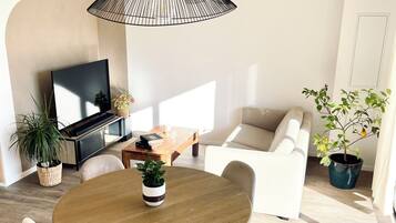 Ein modernes Wohnzimmer mit einem runden Holzesstisch, weißen Stühlen, einer schwarzen Pendelleuchte, einem Flachbildfernseher, einer Couch und Topfpflanzen.