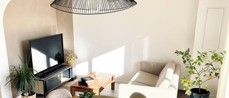 A modern living room with a round wooden dining table, white chairs, a black pendant light, a flat-screen TV, a sofa, and potted plants.