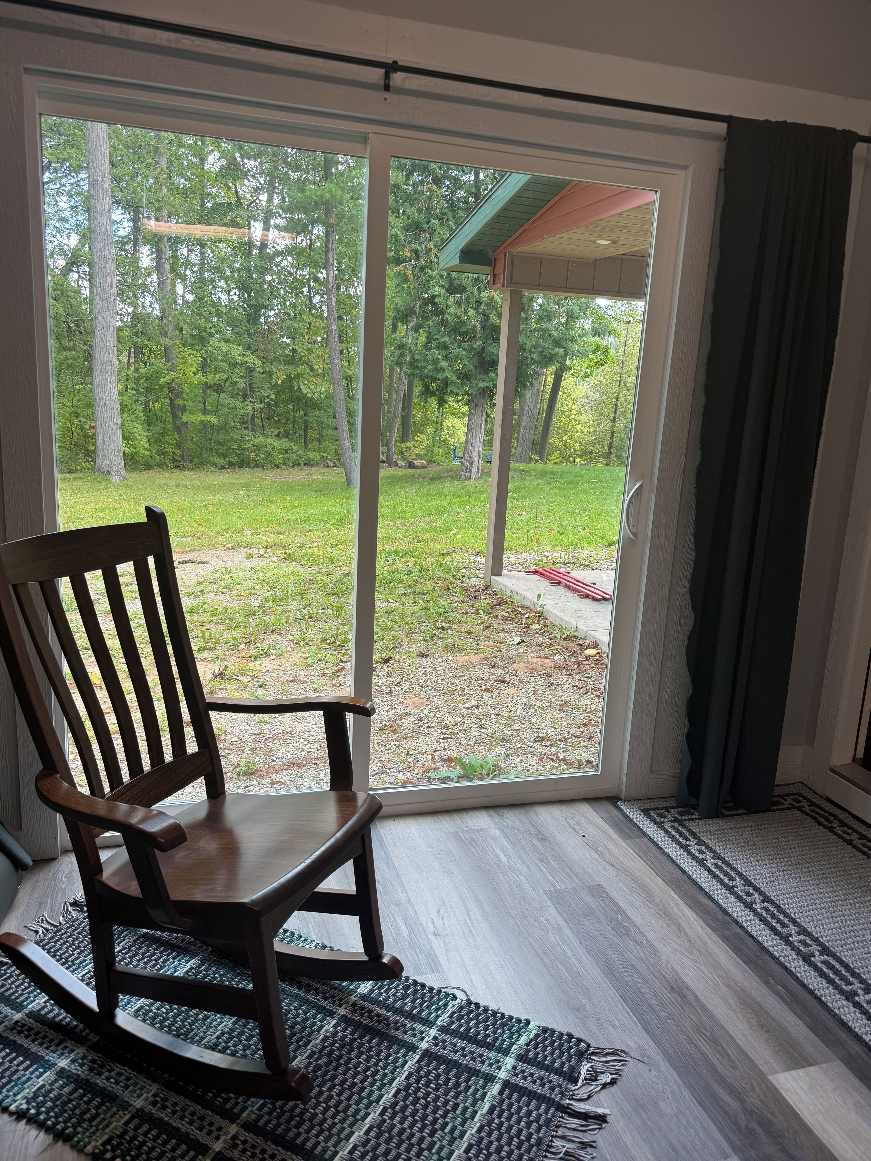 A wooden rocking chair in front of a sliding glass door leading to a covered outdoor area with a view of a grassy yard and trees.