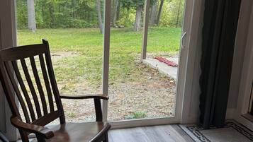 A wooden rocking chair in front of a sliding glass door leading to a covered outdoor area with a view of a grassy yard and trees.