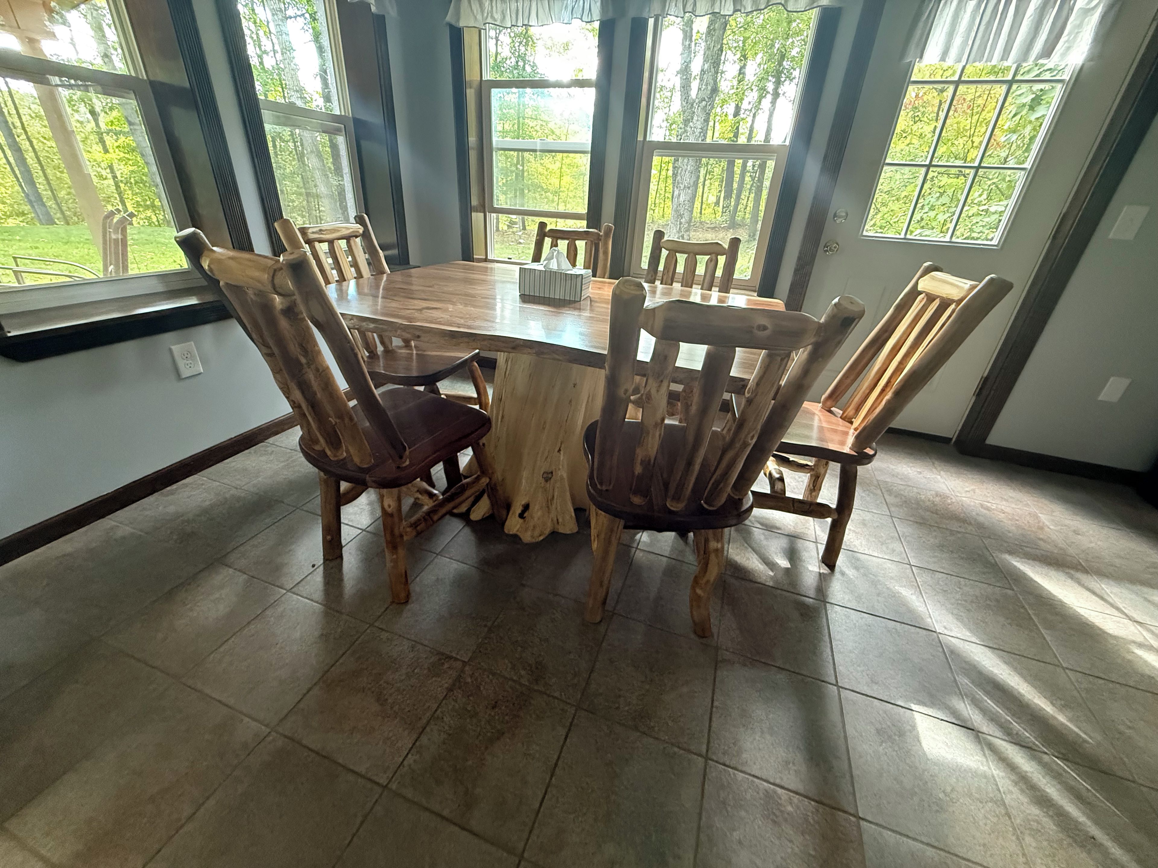 A dining area with a round wooden table surrounded by six chairs, large windows with a view of greenery, and a tiled floor.