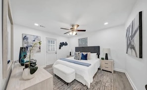 A bedroom with a bed, bedside tables, a ceiling fan, artwork on the walls, and a window with blinds.