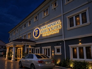 L’entrée d’un hôtel, avec une enseigne indiquant « Posh Imperial Hotel & Suites » et une voiture garée.