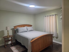 Ein Schlafzimmer mit einem hölzernen Bett, einem Nachttisch mit Lampe, einem Fenster mit Vorhängen und einem kleinen Teppich.