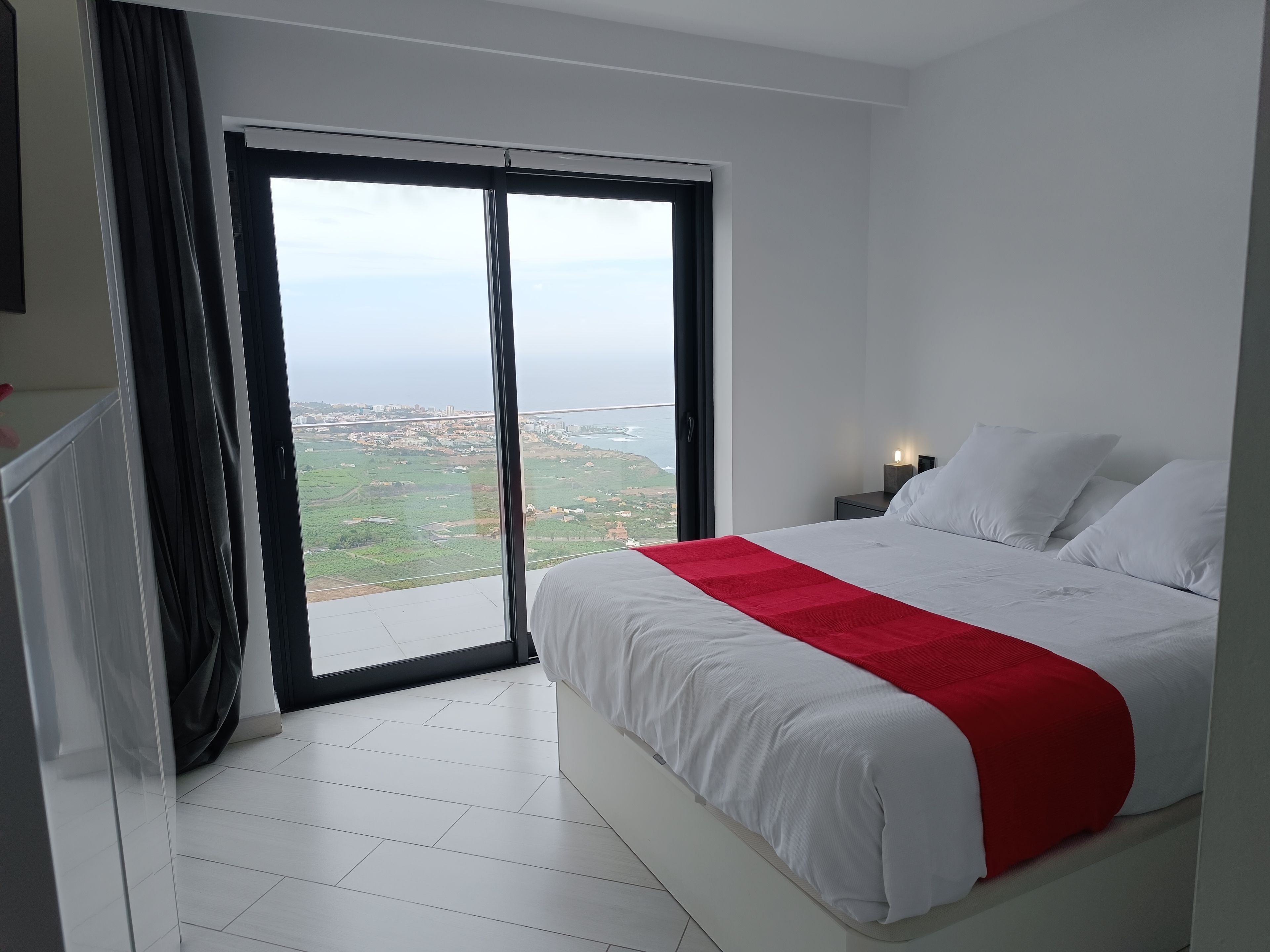 A bedroom with a large bed, a nightstand, a red throw, and a view of a coastal landscape.
