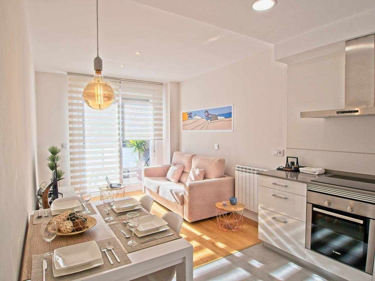 Una cocina moderna con horno empotrado, un comedor con mesa lista para dos personas, y una sala de estar con sofá y un cuadro en la pared.
