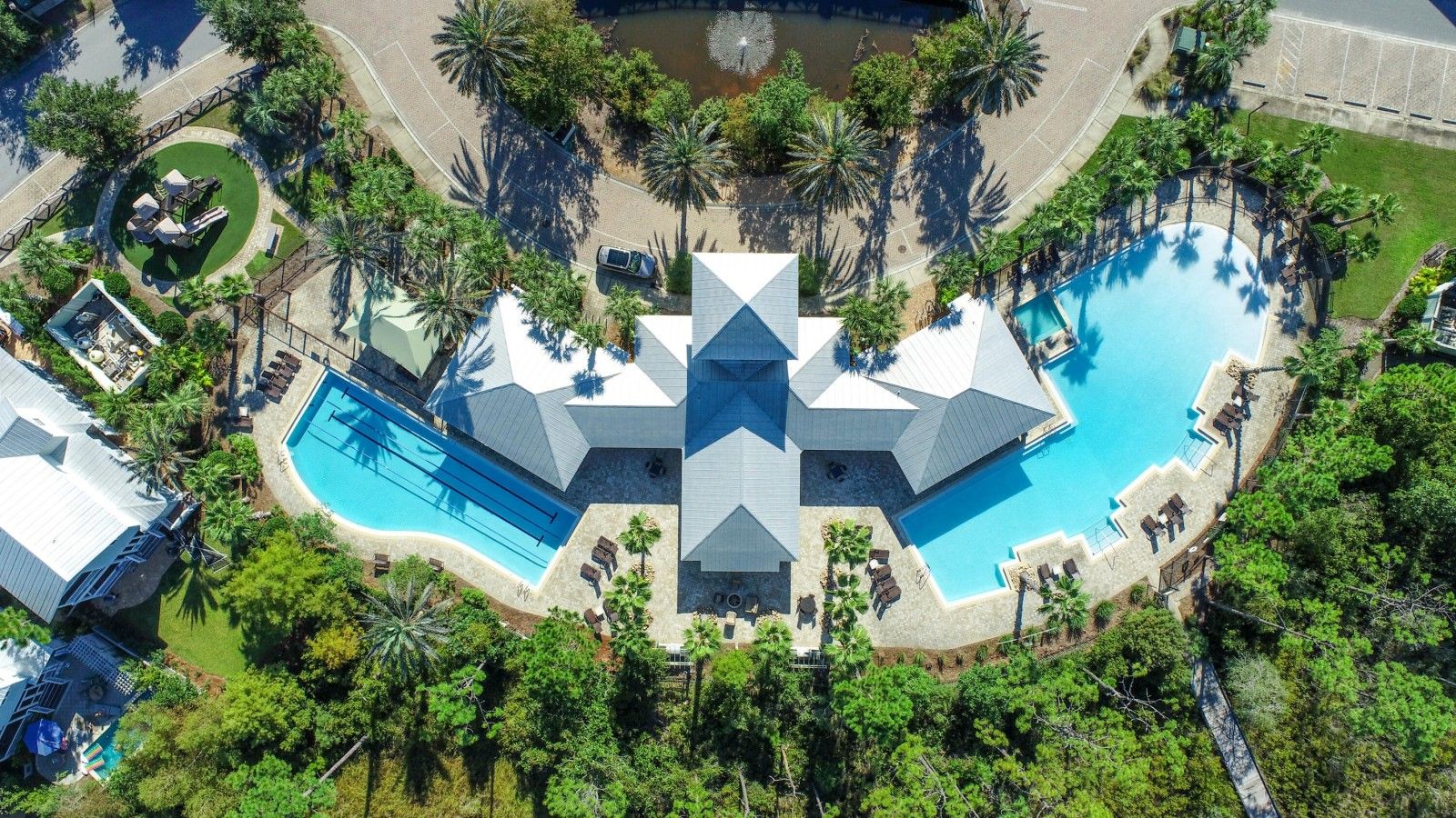 An aerial view of a resort with multiple swimming pools, palm trees, and a central building with a unique geometric roof design.