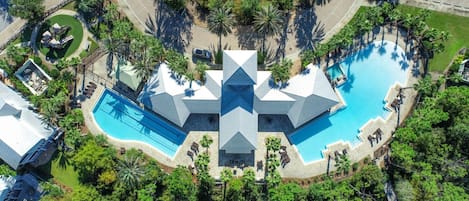 An aerial view of a resort with multiple swimming pools, palm trees, and a central building with a unique geometric roof design.