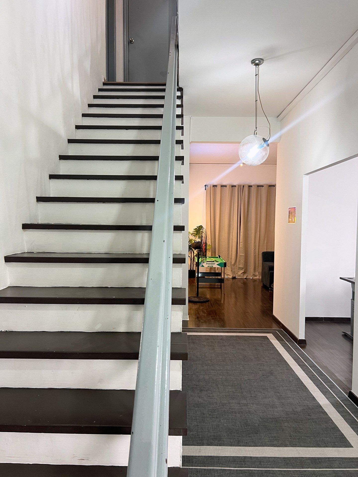 A staircase with alternating black and white steps, a hallway with a ceiling light fixture, and a room with a plant and a Snooker table.