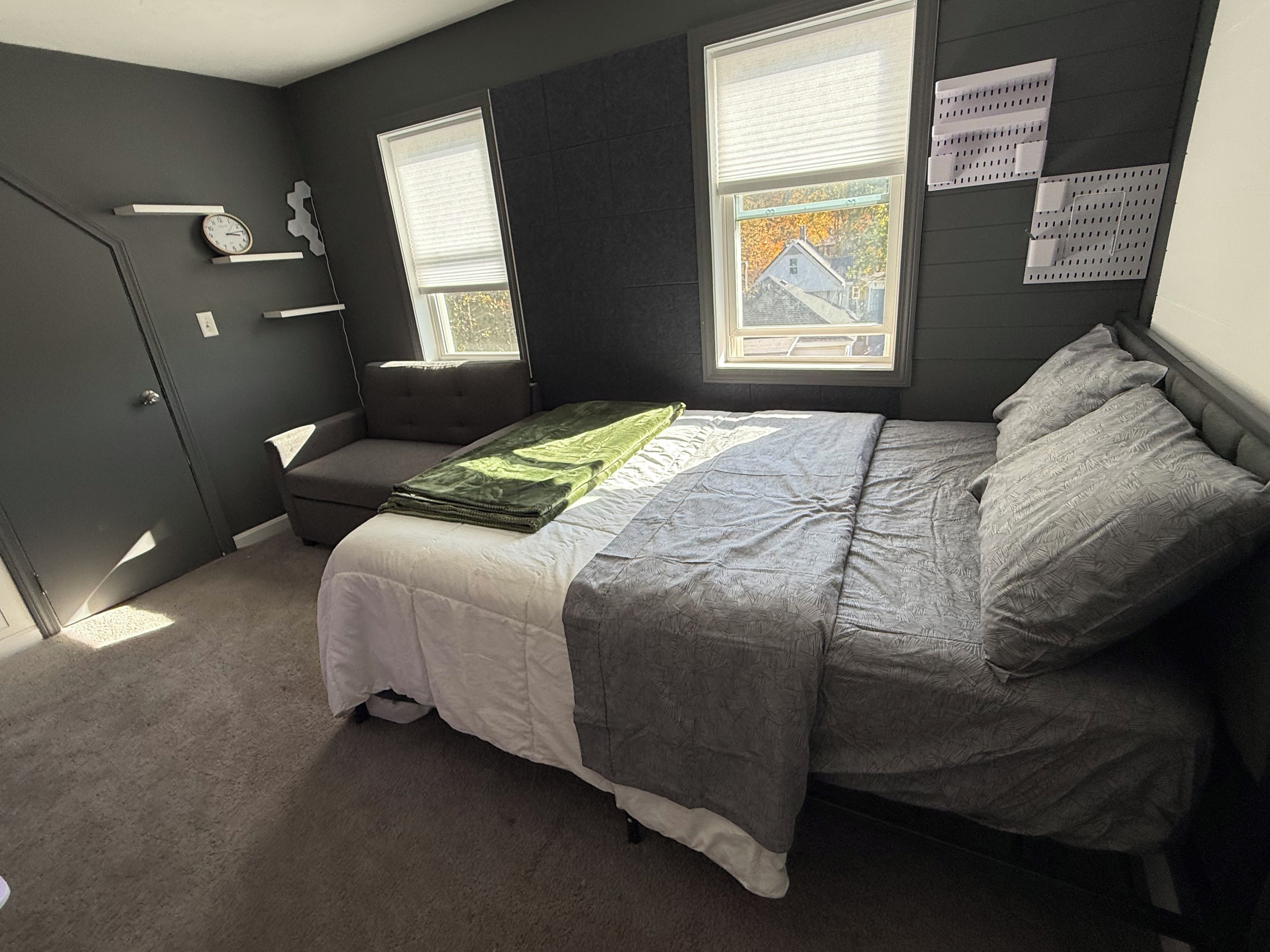 Quarto com cama, um banquinho, janela e prateleira fixada na parede.