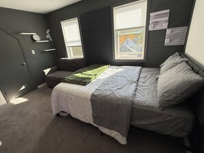 Quarto com cama, um banquinho, janela e prateleira fixada na parede.