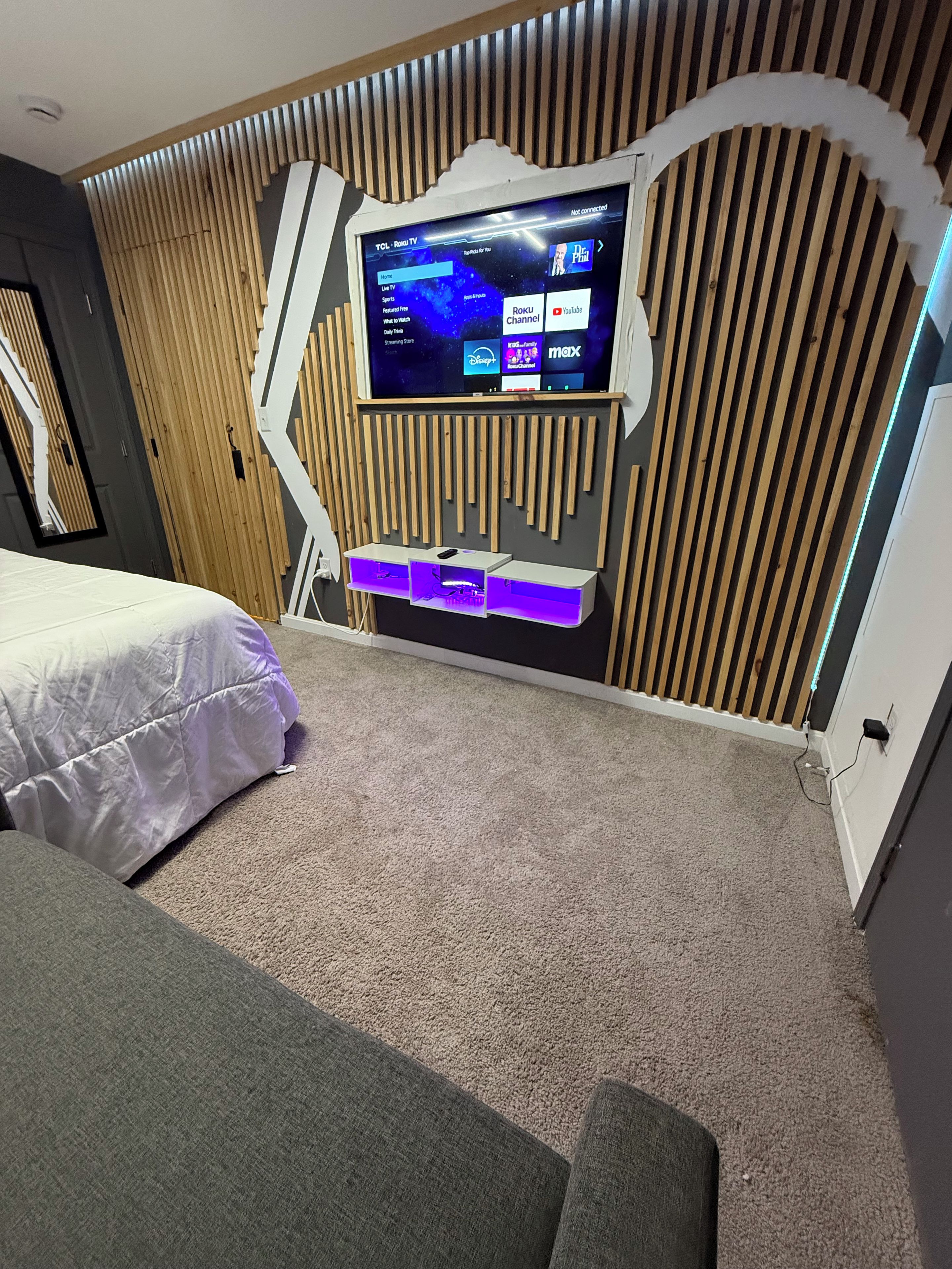 A modern hotel room with a large flat-screen TV mounted on a wooden wall, a bed with a white comforter, and a carpeted floor.
