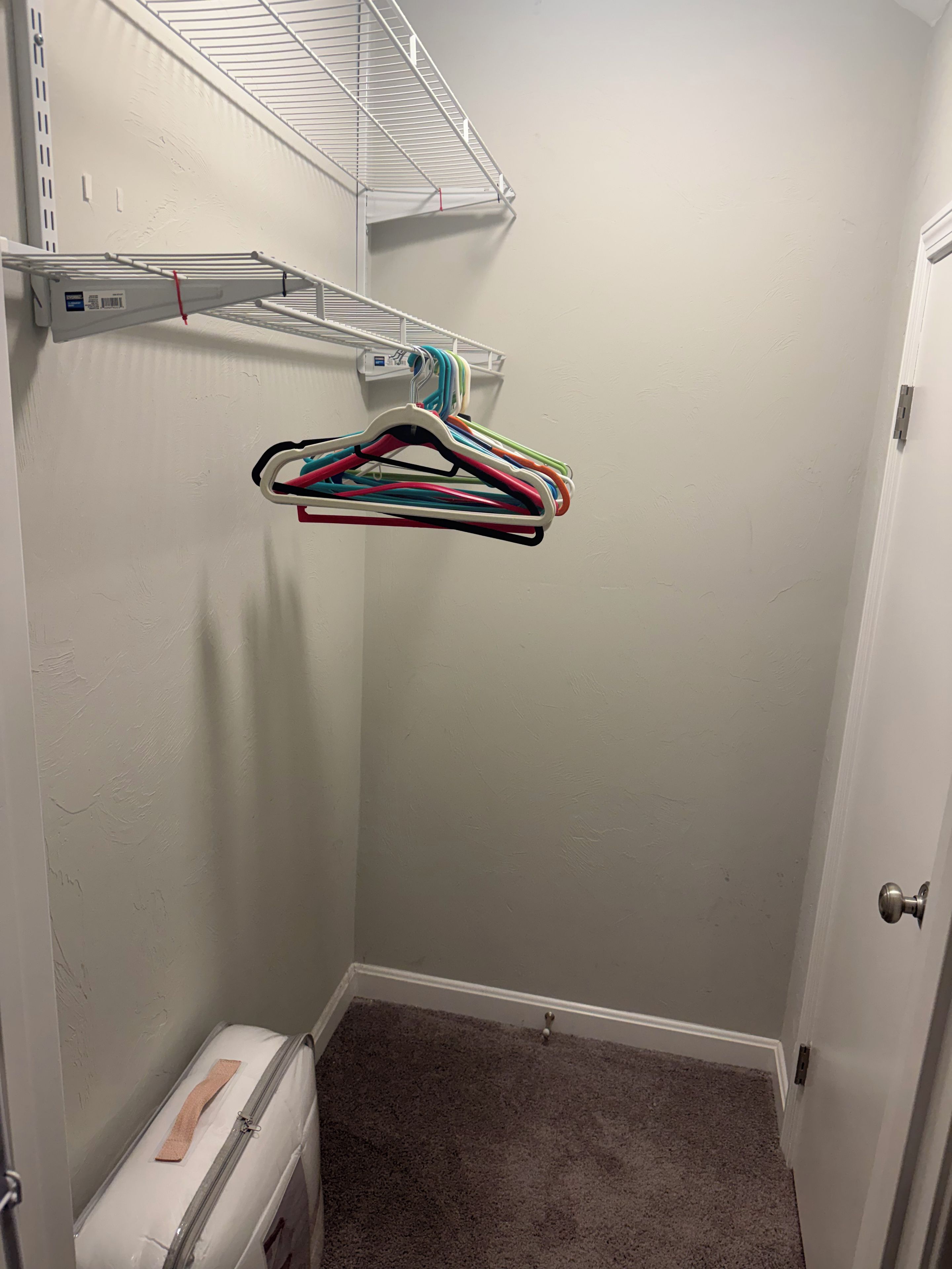 A corner room with a white door, a small white storage unit, and a hanging clothes rack with multiple colorful hangers.