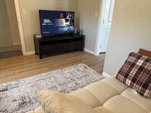 Una sala de estar con un televisor mostrando una película, un mueble de entretenimiento de madera oscura, un sofá color beige y una alfombra estampada.