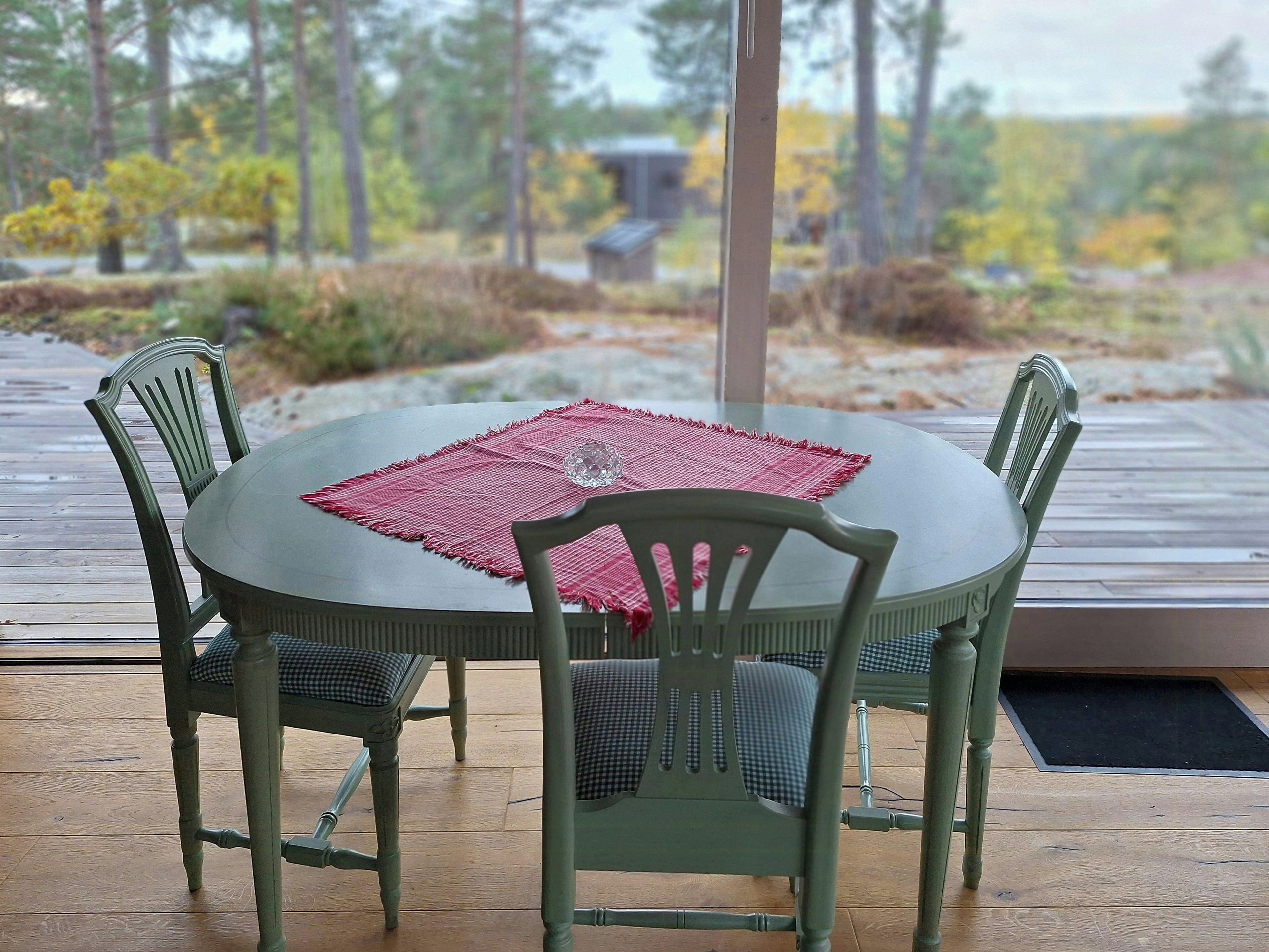 Une table ronde surmontée d’un set de table rouge, entourée de quatre chaises dotées de coussins à carreaux, est placée sur une terrasse en bois offrant une vue sur des arbres et un bâtiment en arrière-plan.